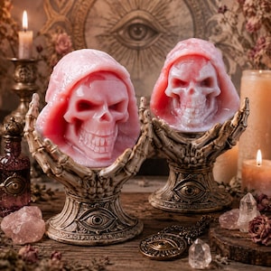 May include: Two pink skull-shaped candles with hooded details, each resting in a decorative beige skeletal hand pedestal base. The bases feature an eye motif at the center of the stand. The candles are crafted with realistic facial features.