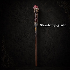 May include: A slender wand featuring a dark brown handle and a textured, metallic gold-toned top section adorned with a rough-cut, pinkish-red Strawberry Quartz crystal at the tip. The text Strawberry Quartz is displayed to the right of the wand.