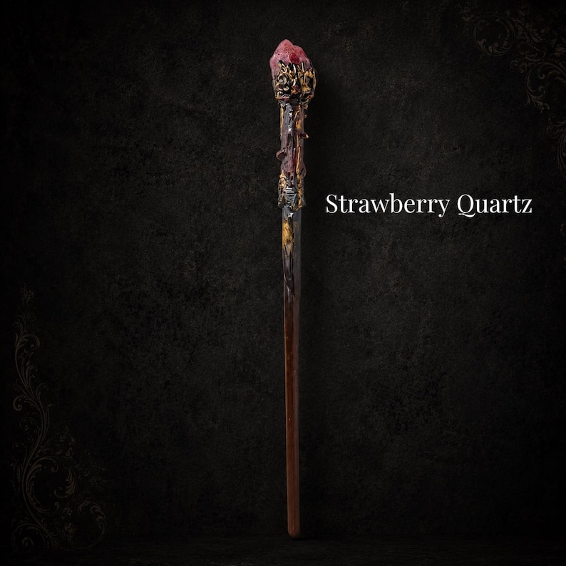 May include: A slender wand featuring a dark brown handle and a textured, metallic gold-toned top section adorned with a rough-cut, pinkish-red Strawberry Quartz crystal at the tip. The text Strawberry Quartz is displayed to the right of the wand.