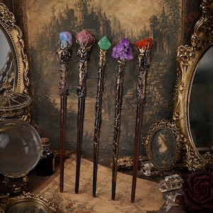 May include: Five wands with dark brown, textured handles and metallic gold accents. Each wand is topped with a unique crystal: a blue stone, a pink stone, a green stone, a cluster of purple amethyst, and a single orange stone, all held by metal claws.