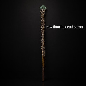 May include: A long, slender wand featuring a dark brown, textured handle wrapped in metallic gold wire vines. The top is crowned with a translucent, green raw fluorite octahedron crystal. The text "raw fluorite octahedron" appears to the right.