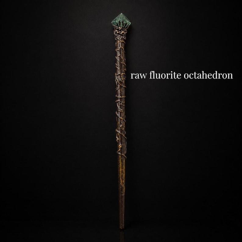 May include: A long, slender wand featuring a dark brown, textured handle wrapped in metallic gold wire vines. The top is crowned with a translucent, green raw fluorite octahedron crystal. The text "raw fluorite octahedron" appears to the right.