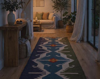 Op de afbeelding: Een rechthoekig vloerkleed met een geometrisch patroon in blauwe, groene, witte en rode tinten. Het kleed heeft een centraal ruitmotief, een gekartelde rand en korte franjes aan beide uiteinden, liggend op een houten vloer.