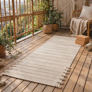May include: A rectangular off-white woven runner rug featuring a textured striped pattern and fringed edges on both ends. The rug is positioned on a wooden deck floor, surrounded by potted plants, a rolled yoga mat, and two stacked yoga blocks.