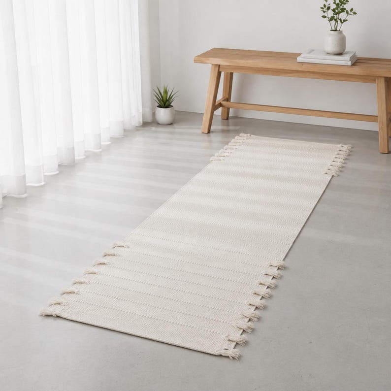 May include: A long, rectangular off-white runner rug featuring a textured woven pattern and decorative fringe detailing along both short ends. The rug is positioned flat on a smooth, light-colored floor surface.