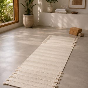 May include: A rectangular off-white woven runner rug featuring a textured striped pattern and decorative knotted fringe tassels along both of the shorter ends. The rug is constructed with a dense, uniform weave and a neutral, light beige color.