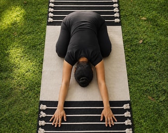 May include: A rectangular yoga mat featuring a central beige panel flanked by two black sections. The black ends are decorated with horizontal white rope fringe details that extend across the width of the mat, creating a textured contrast.