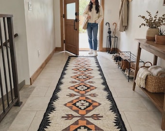 Op de afbeelding: Een lang rechthoekig vloerkleed met een crèmekleurige basis en een herhalend geometrisch patroon in zwart, donkergrijs en oranje. Het kleed heeft een dikke zwarte rand en is afgewerkt met korte, geknoopte grijze franjes aan beide uiteinden.