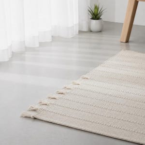 May include: A rectangular off-white area rug featuring a textured, ribbed pattern across its surface. The rug is finished with short, knotted fringe tassels along the edge, providing a tactile detail to the woven fabric construction.