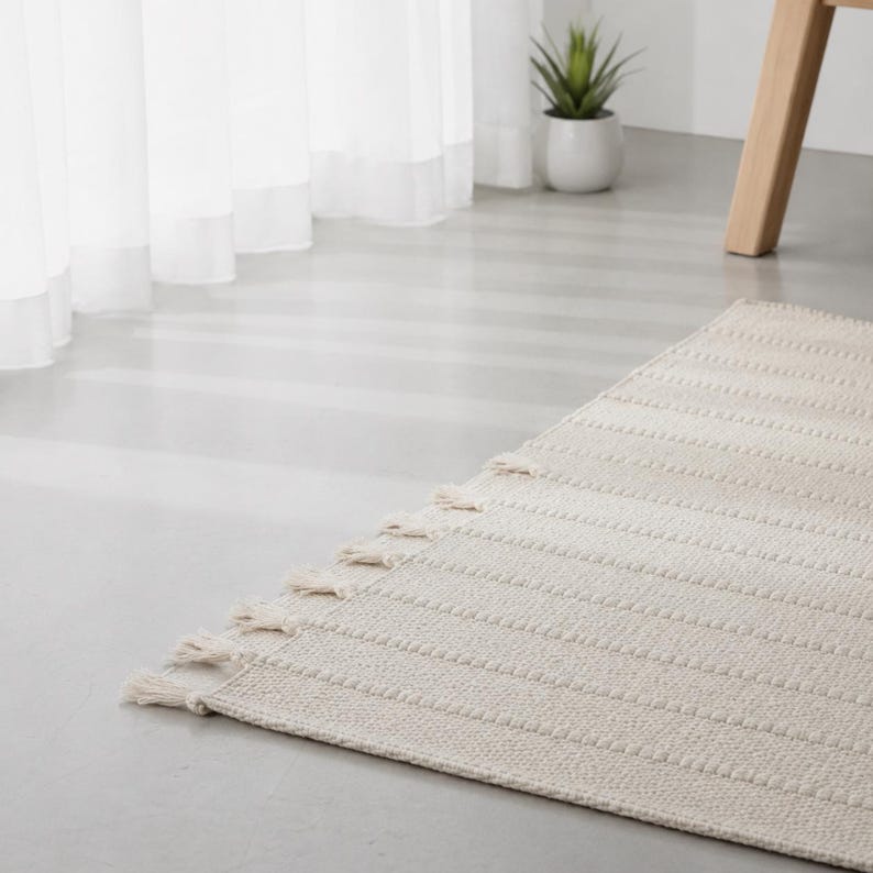 May include: A rectangular off-white area rug featuring a textured, ribbed pattern across its surface. The rug is finished with short, knotted fringe tassels along the edge, providing a tactile detail to the woven fabric construction.