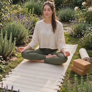 May include: A cream-colored yoga mat with fringe detailing along the sides, accompanied by two rectangular brown foam yoga blocks and a rolled-up cream yoga mat, all positioned on a grassy surface with lavender and greenery.
