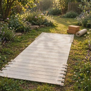 May include: A rectangular off-white woven mat featuring a textured striped pattern and decorative fringe tassels along the shorter edges, accompanied by two rectangular cork blocks and a rolled-up mat in the background.
