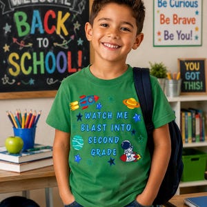 Puede incluir: Una camiseta verde de manga corta con un gráfico de temática espacial que incluye un cohete, un planeta y un astronauta. El texto "WATCH ME BLAST INTO SECOND GRADE" aparece en letras azules, rodeado de pequeñas estrellas en varios tonos de azul.