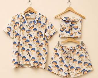 May include: A short-sleeve button-down pajama shirt and matching drawstring shorts featuring an all-over print of blue-capped mushrooms with tan stems on a cream background. The set includes a folded fabric piece hanging on a wooden hanger.
