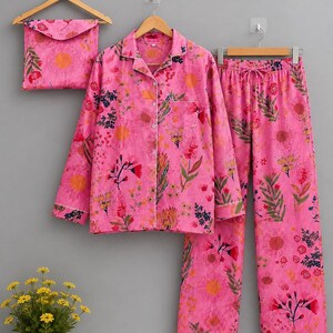 May include: A bright pink pajama set featuring a long-sleeved button-down shirt with a collar and a chest pocket, paired with matching drawstring pants. Both pieces display a vibrant floral print with orange, red, and green botanical motifs.