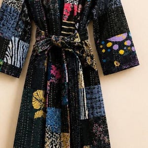 May include: A long-sleeved robe featuring a patchwork design of various dark-toned fabrics with multicolored floral, geometric, and abstract patterns. The garment includes a matching tie belt at the waist and visible white running stitch details.