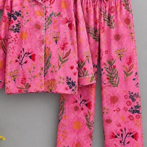 May include: A bright pink pajama set featuring a long-sleeved button-down shirt with a collar and a chest pocket, paired with matching long pants that have an elastic drawstring waistband, all decorated with a colorful floral and botanical print.