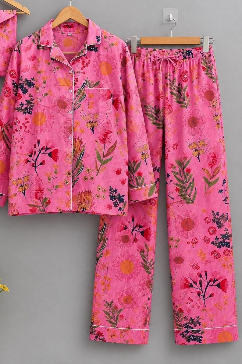 May include: A bright pink pajama set featuring a long-sleeved button-down shirt with a collar and a chest pocket, paired with matching long pants that have an elastic drawstring waistband, all decorated with a colorful floral and botanical print.