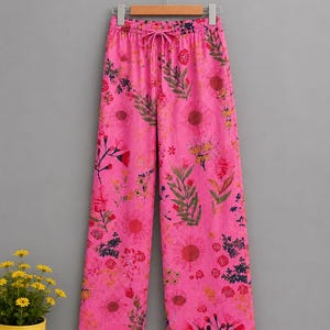 May include: Bright pink wide-leg trousers featuring an all-over floral print in shades of red, orange, yellow, and dark green. The pants have an elasticized waistband with a matching drawstring tie and a thin white horizontal stripe at the hem.