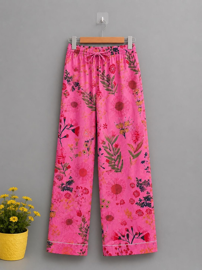 May include: Bright pink wide-leg trousers featuring an all-over floral print in shades of red, orange, yellow, and dark green. The pants have an elasticized waistband with a matching drawstring tie and a thin white horizontal stripe at the hem.
