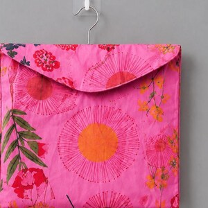 May include: A square fabric storage pouch in a vibrant pink hue, featuring a printed pattern of large orange and pink circular floral motifs, green leaves, and small yellow blossoms. The pouch has a triangular flap and hangs from a metal hook.
