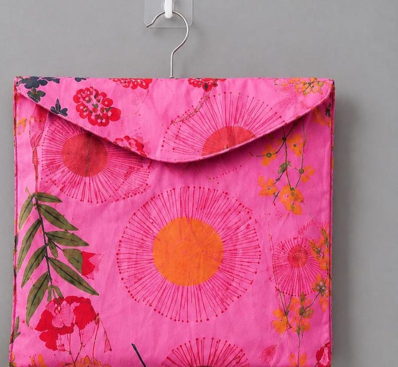 May include: A square fabric storage pouch in a vibrant pink hue, featuring a printed pattern of large orange and pink circular floral motifs, green leaves, and small yellow blossoms. The pouch has a triangular flap and hangs from a metal hook.