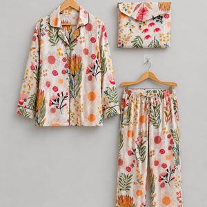 May include: A long-sleeve button-down pajama shirt, matching drawstring pajama pants, and a folded fabric pouch, all featuring a cream-colored base with a repeating pattern of orange suns, red flowers, and green foliage with thin black piping.
