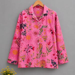 May include: A long-sleeved button-down pajama shirt in bright pink featuring a colorful floral and botanical print. The shirt includes a notched collar, a single chest pocket, and white piping trim along the collar, front placket, and sleeve cuffs.