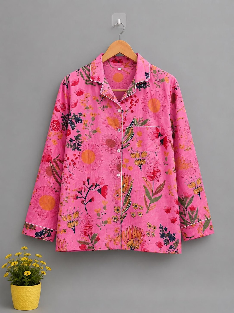 May include: A long-sleeved button-down pajama shirt in bright pink featuring a colorful floral and botanical print. The shirt includes a notched collar, a single chest pocket, and white piping trim along the collar, front placket, and sleeve cuffs.