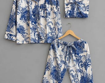 May include: A pajama set consisting of a long-sleeved button-down shirt with a collar and a chest pocket, a pair of drawstring pants, and a folded square fabric piece, all featuring a blue and white toile print of tropical trees and birds.