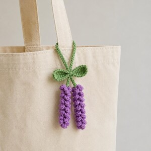 Peut inclure: Une breloque fleur de lavande au crochet violet et vert suspendue à la anse d'un sac fourre-tout en toile beige. La breloque présente deux tiges de fleurs violettes texturées attachées à une tige verte avec deux petites feuilles vertes.