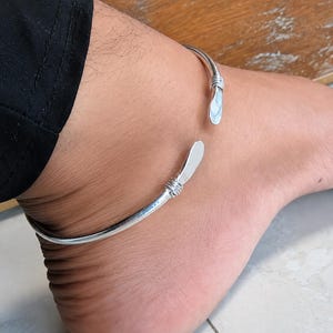 May include: A silver-toned open cuff anklet featuring a smooth, polished finish. The design includes two flat, paddle-shaped terminals at each end, secured by decorative wire-wrapped detailing just before the flattened tips of the metal band.