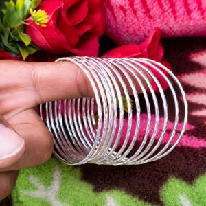 May include: A set of multiple thin, silver-colored metal bangles with a hammered, textured finish. The circular bracelets are stacked together, creating a layered appearance with reflective surfaces that catch the light.