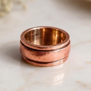 May include: A wide copper-coloured metal ring featuring a smooth, polished central band flanked by two thinner, raised ridges along the top and bottom edges. The ring has a warm, metallic finish with subtle variations in tone across its surface.
