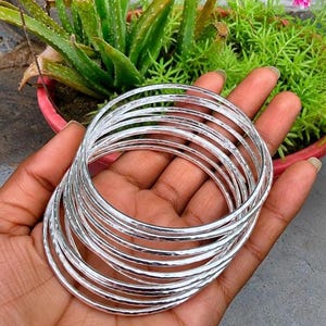 Può includere: Un set di sette braccialetti circolari sottili in argento con una finitura martellata e testurizzata. Ogni braccialetto presenta una superficie metallica riflettente che cattura la luce, creando un aspetto sovrapposto quando indossati insieme.