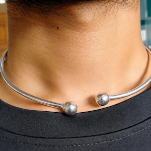 May include: A polished silver-tone metal choker necklace featuring a rigid, open-ended circular band design. The ends of the necklace are finished with two smooth, spherical ball terminals that face each other at the front center of the neck.