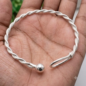 Può includere: Un bracciale a fascia color argento caratterizzato da un design a corda intrecciata. Il bracciale ha una struttura aperta con una sfera liscia e lucida a un'estremità e un semplice anello arrotondato all'altra per una chiusura sicura.