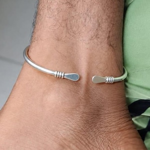 May include: A silver-toned open cuff anklet featuring a smooth, rounded band. Each end of the anklet terminates in a flattened, paddle-shaped tip, accented by three distinct horizontal grooves just before the flat terminal point.