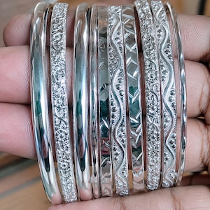 Può includere: Un set di otto braccialetti rigidi color argento caratterizzati da una varietà di texture e motivi. La collezione include fasce lisce lucide, intricate incisioni floreali, linee ondulate punteggiate e intagli geometrici a forma di diamante.