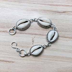 Peut inclure: Un bracelet argenté composé de quatre coquillages cauris blancs, chacun enchâssé dans un cadre métallique perlé décoratif. Le bracelet est relié par des maillons en métal argenté et comprend un fermoir à crochet et un anneau circulaire.