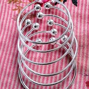 Może przedstawiać: Stos pięciu polerowanych bransoletek typu bangle w kolorze srebrnym, z których każda ma okrągły, otwarty kształt z gładką kulką na obu końcach. Bransoletki są ułożone w pionowej kolumnie, eksponując swoją błyszczącą i odblaskową powierzchnię.