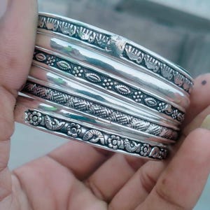 Puede incluir: Una pila de cuatro brazaletes de metal plateado con distintos patrones en relieve. Los diseños incluyen una fila de pequeños elefantes, un motivo floral y de cuentas, una textura de cuadrícula y una banda decorada con flores y hojas alternas.