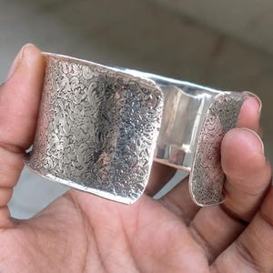 Può includere: Un ampio bracciale a fascia in argento caratterizzato da un intricato motivo floreale e a volute in rilievo su tutta la superficie esterna. Il metallo presenta una finitura lucida e riflettente con una texture che esalta le incisioni.