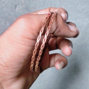 Può includere: Due braccialetti color rame caratterizzati da un design intrecciato a forma di corda. I bracciali presentano una finitura metallica e un motivo ripetitivo continuo lungo l'intera circonferenza di ogni fascia circolare, offrendo un aspetto strutturato uniforme.