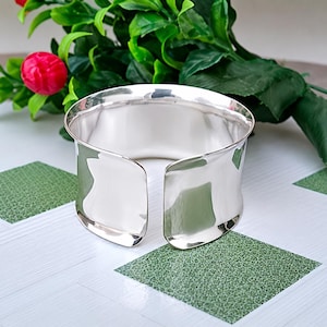 Può includere: Un ampio bracciale a fascia in argento lucido con una superficie liscia e riflettente e un design aperto sul retro. Il metallo ha una finitura molto brillante che cattura la luce, creando un aspetto metallico pulito con bordi curvi.