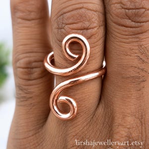 May include: A polished copper-colored ring featuring a bypass design with two open-ended spirals. The metal wire is smooth and reflective, wrapping around the finger. The text tirshajewelleryart.etsy is visible at the bottom of the image.