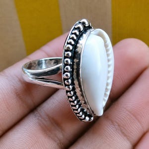 May include: A silver ring featuring a central white cowrie shell set within a decorative, beaded metal frame. The band of the ring is split into two distinct sections where it connects to the oval-shaped setting, showcasing a polished finish.