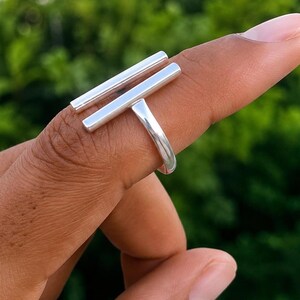 Può includere: Un anello in metallo argentato lucido caratterizzato da due barre orizzontali parallele che si estendono sul dito. La fascia si collega al centro della barra inferiore, creando un design geometrico pulito con una finitura metallica liscia.