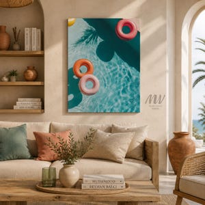 May include: A rectangular wall art print featuring an aerial view of a turquoise swimming pool with three inflatable pool rings in pink, orange, and yellow floating on the water surface, casting dark shadows on the pool floor below.