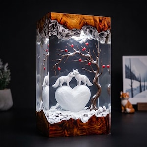 May include: A rectangular resin lamp featuring a natural wood base and top. Inside, two white fox figurines stand on a white heart-shaped mound beneath a brown tree with red berries. The scene is set against a snowy landscape with clear resin.
